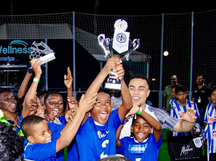 Young players in blue jerseys celebrating with a trophy at the Sergiño Dest Court